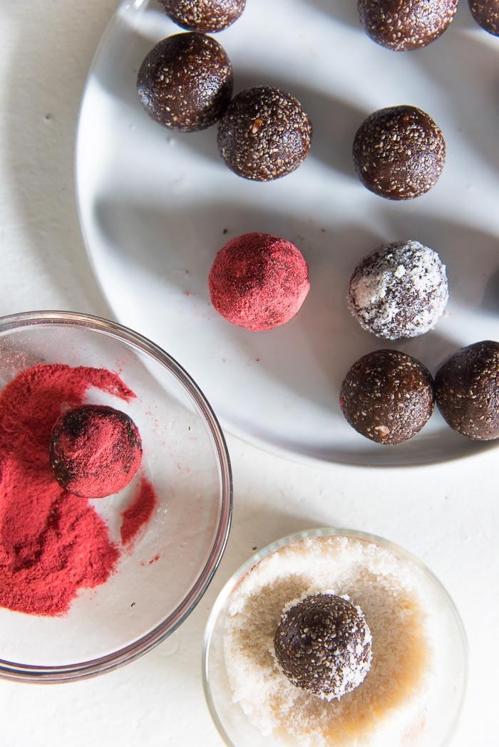 Red, White and Brown colored Fudgy Chocolate Chia bliss balls on a white plate, being coated with coconut (in one bowl), and freeze dried strawberry powder (in another bowl). 