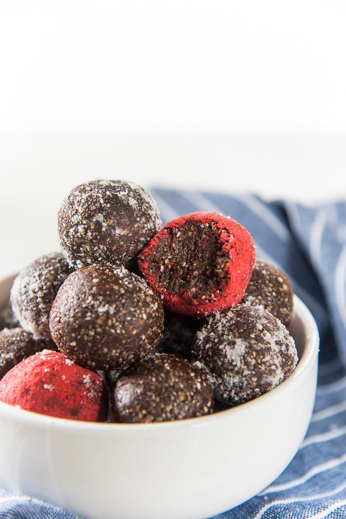Fudgy Chocolate Chia Balls piled up in a white bowl. One strawberry coated chocolate bliss ball has a bite taken out to show how fudgy the center of the bliss balls are. 