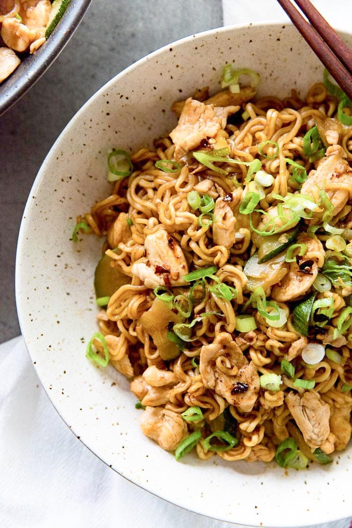 Une photo aérienne des nouilles sautées au poulet Ramen 15 minutes qui est servie dans un bol avec des courgettes, du poulet, des oignons verts et de l'huile de piment de sésame.