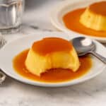 Two plates of flan with caramel sauce on a marble surface, one partially eaten with a spoon beside it.