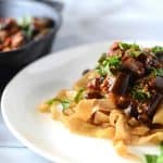 A white plate with pasta topped with a generous serving of roasted eggplant.