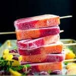Stack of peach melba popsicles (paletas) on a sheet pan with peach slices and raspberries next to it.