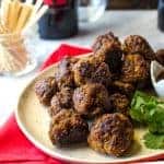 A plate of meatballs on a beige dish, garnished with fresh parsley, and a glass with toothpicks and red napkin in the background.