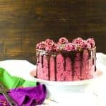 A vibrant pink cake with chocolate drizzle and colorful sprinkles sits on a white cake stand.