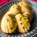 A plate of six golden-brown madeleines, sprinkled with passion fruit glaze and seeds, arranged on a silver platter.