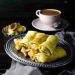 Metal tray of sweet coconut filled crepes (pani pol) with a cup of tea in the background.