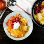 Blood orange crepes suzette on a plate, served with blood orange slices, raspberries, and whipped cream.