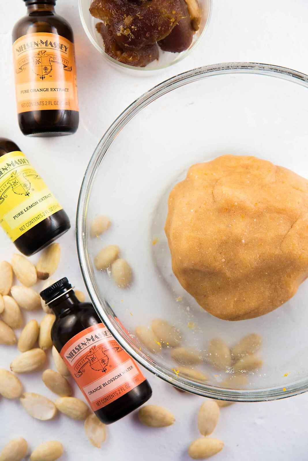 Almond paste in a big glass bowl with small bottles of flavoring next to it.