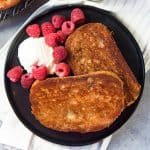 Two slices of caramelized cinnamon toast on a black plate served with a dollop of whipped cream and fresh raspberries.