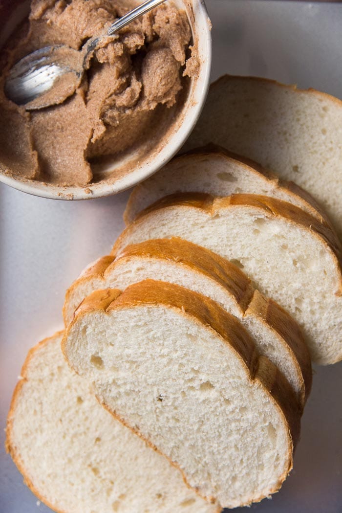 An overhead image of sliced country bread on a white cutting board, next to a bowl with cinnamon butter.