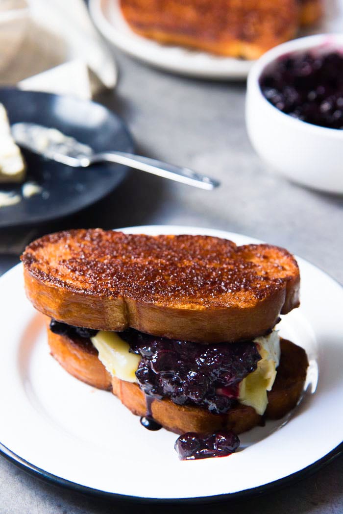 A close up of Two slices of buttery cinnamon toast as a sandwich with melted brie and blueberry compote spilling out from the middle, on a white plate.