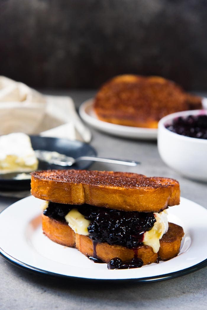 Two slices of buttery cinnamon toast as a sandwich with melted brie and blueberry compote in the middle, on a white plate.