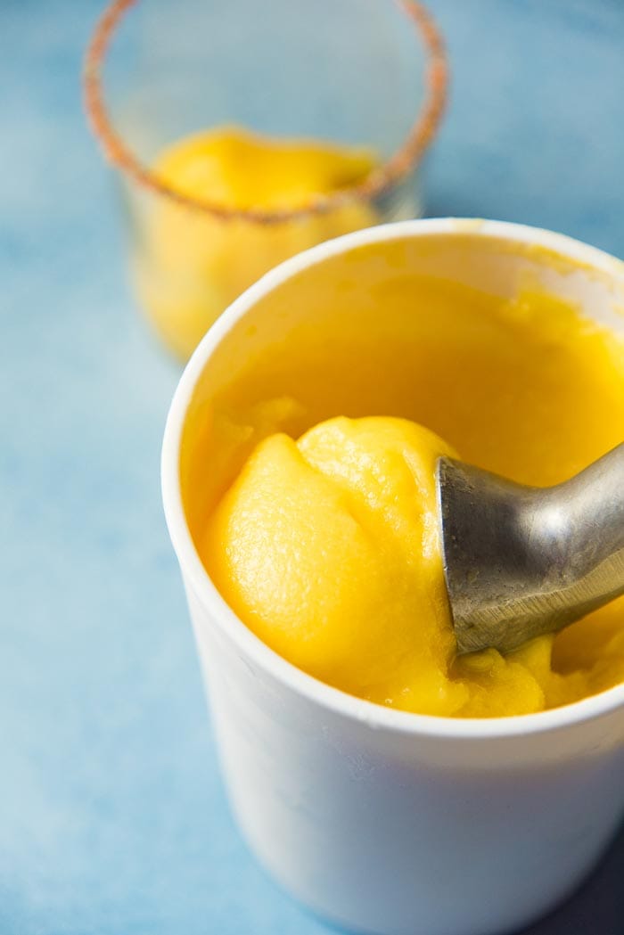 This sorbet like Frozen Mango Margarita in an ice cream scoop in the ice ream container. A Tajín rimmed glass in the background. 