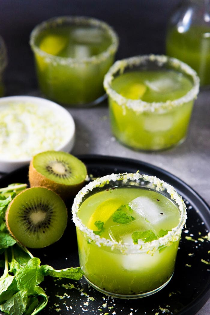 One Mint kiwi Margarita on a black plate next to one sliced kiwi, and 2 more mint kiwi margaritas in the background, on a gray table top. 