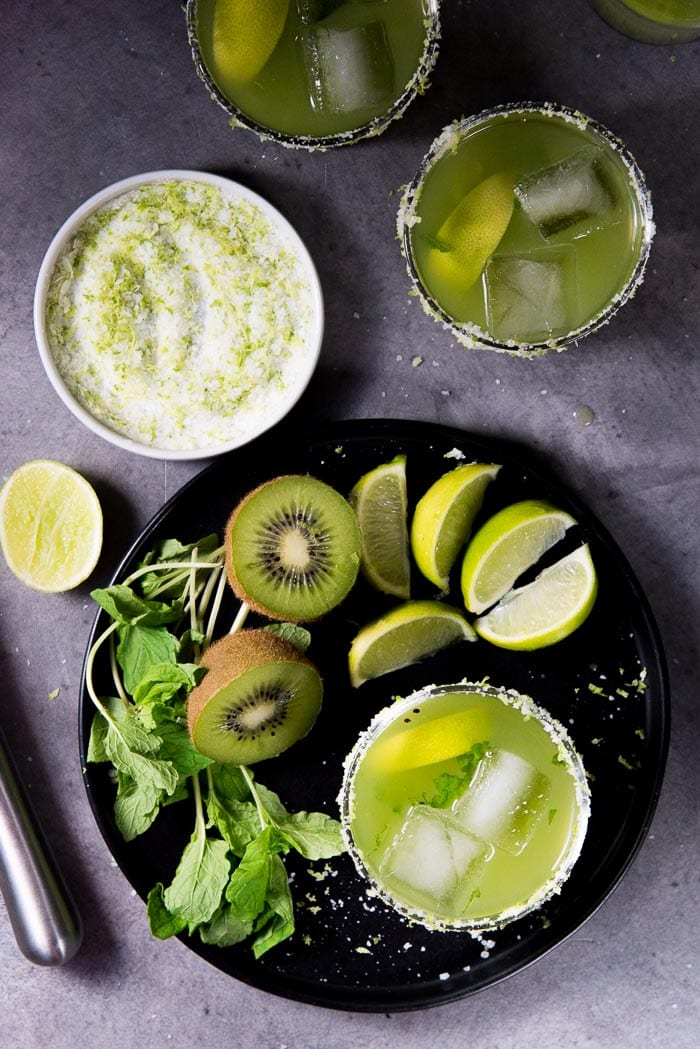 An overhead view of Kiwi Mint Margarita. One cocktail on a black plate next to a bunch of mint, sliced kiwi fruits and lime wedges. A small plate of lime salt and 2 more cocktails in the background. 