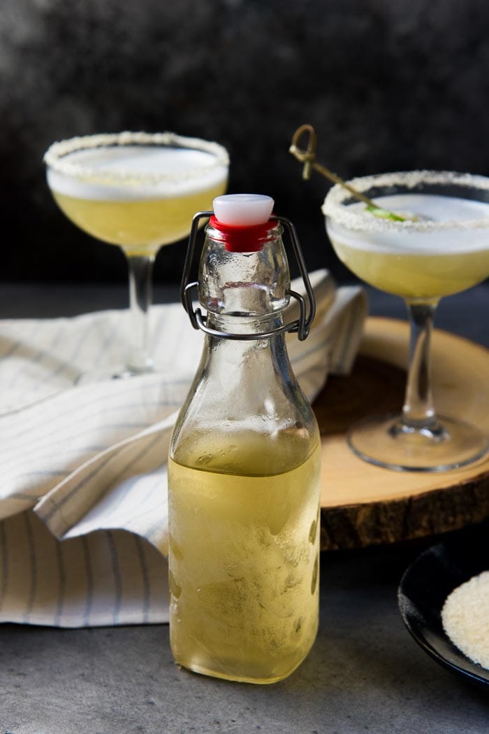 The bottle of chilled smoked tequila, with an air tight stopper. Two Margarita sour cocktails in the background ontop of a wooden stand.