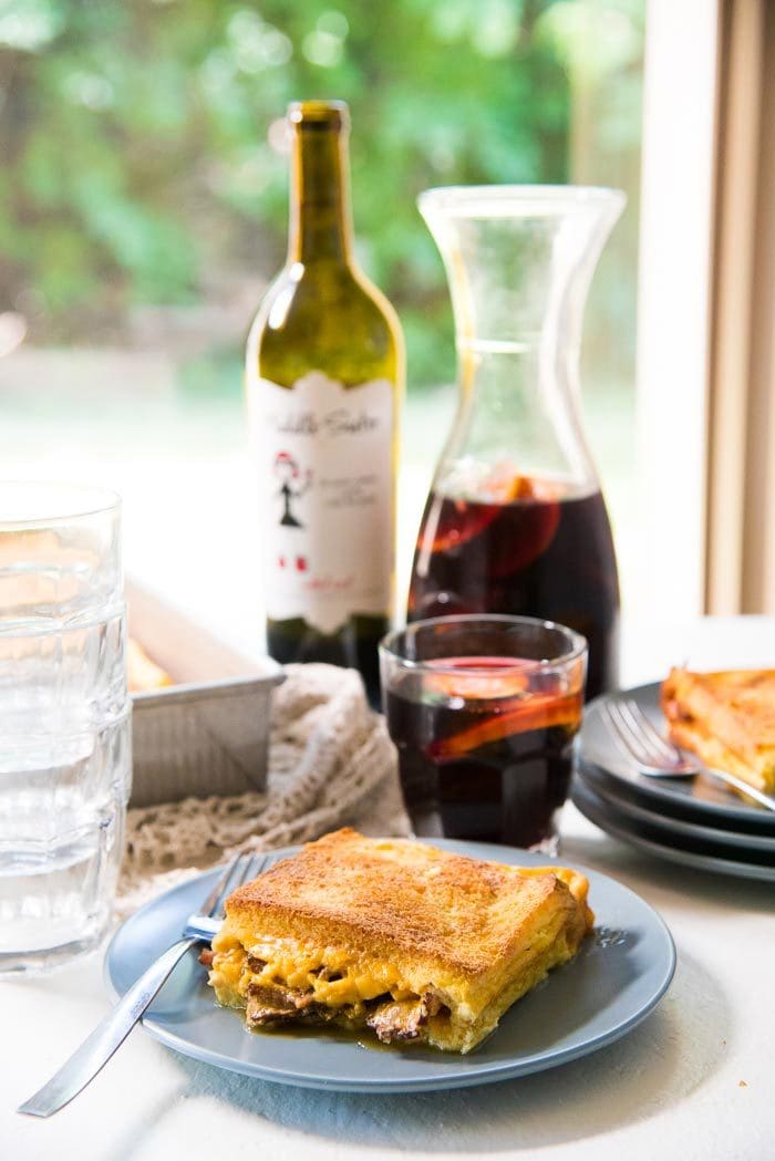A slice of bacon cheese grilled Cheese sandwich casserole on a blue plate, with pieces of bacon and cheese peeking through the edges. With a glass of red wine an elderflower sangria in the background, with a bottle of wine, more plates, and the baking dish in the background. 