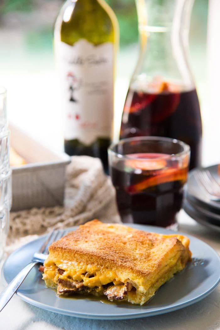 A slice of bacon cheese grilled Cheese sandwich casserole on a blue plate, with pieces of bacon and cheese peeking through the edges. With a glass of red wine an elderflower sangria in the background, with a bottle of wine, more plates, and the baking dish in the background.