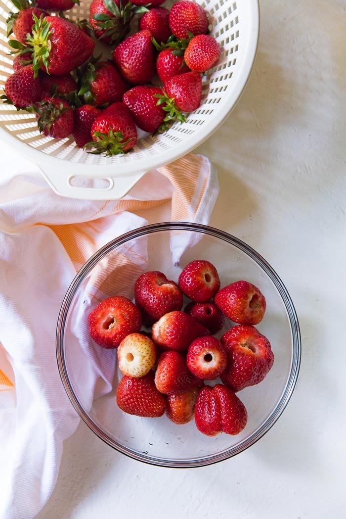 Make sure to hull fresh bright, and sweet strawberries for the recipe. This is an overhead view of a bowl of hulled strawberries, next to unhulled fresh strawberries in a colander.