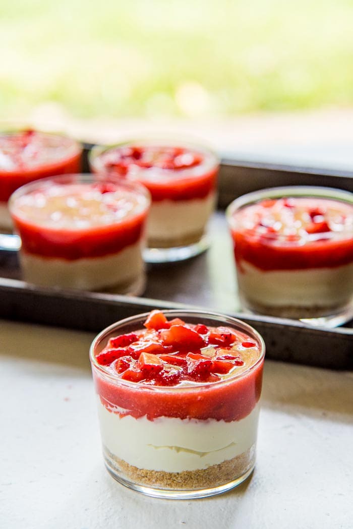 A photo of the serving glasses with the crust, cheesecake and strawberry layers. One dessert is in the foreground, and the others are in the background.