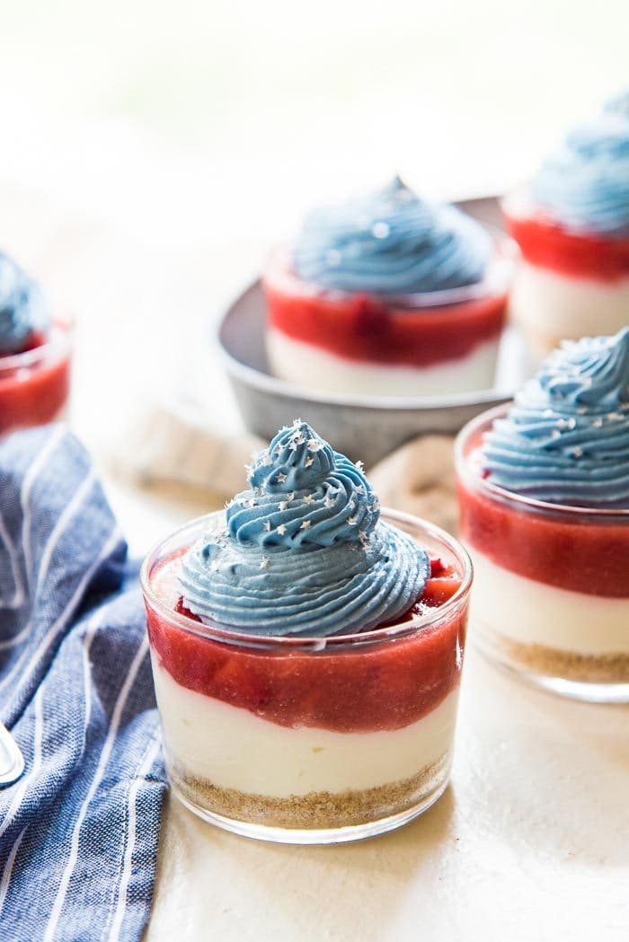 The No Bake Coconut Strawberry Cheesecake Jars on a white table top. The perfect fourth of july dessert for a crowd. One in the foreground, and 3 in the background. Next to a blue and white striped napkin.
