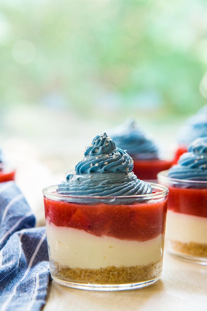 A close up, side on view of the No Bake Coconut Strawberry Cheesecake Jars on a white table top. One focused in the foreground. The bottom layer is a light brown crust, the next layer is a white cheesecake layer that is topped with a fresh strawberry and jello layer and a swirl of blue whipping cream and silver star sprinkles.