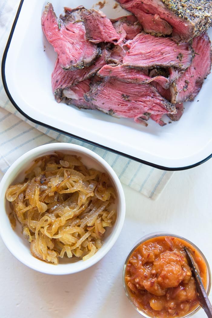 Slow roasted beef thinly sliced for steak sandwiches in an enamel tray. With other steak sandwich fillings like, caramelized onions in one bowl and spicy peach relish in another bowl. 