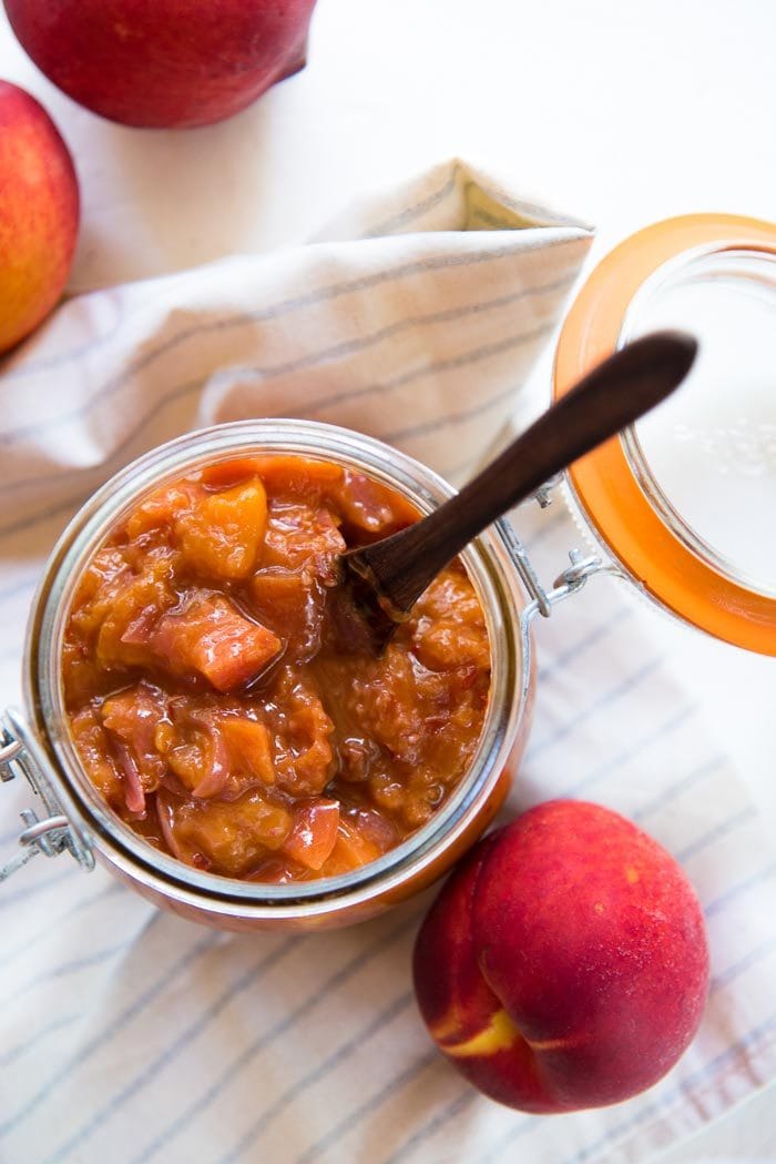 Overhead image of the peach relish in a large ball jar on a striped napkin, next to fresh peaches. 