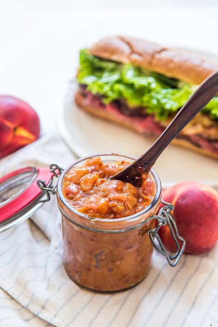 Sweet nd Spicy peach relish in a glass jar, with a wooden serving spoon in it, next to fresh whole peaches. A steak sandwich with peach relish in the background. 
