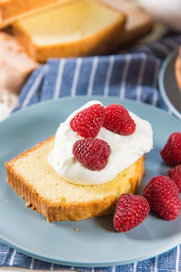 A close up of a slice of pound cake on a blue plate, served with a dollop of whipped cream on top, with fresh raspberries. 