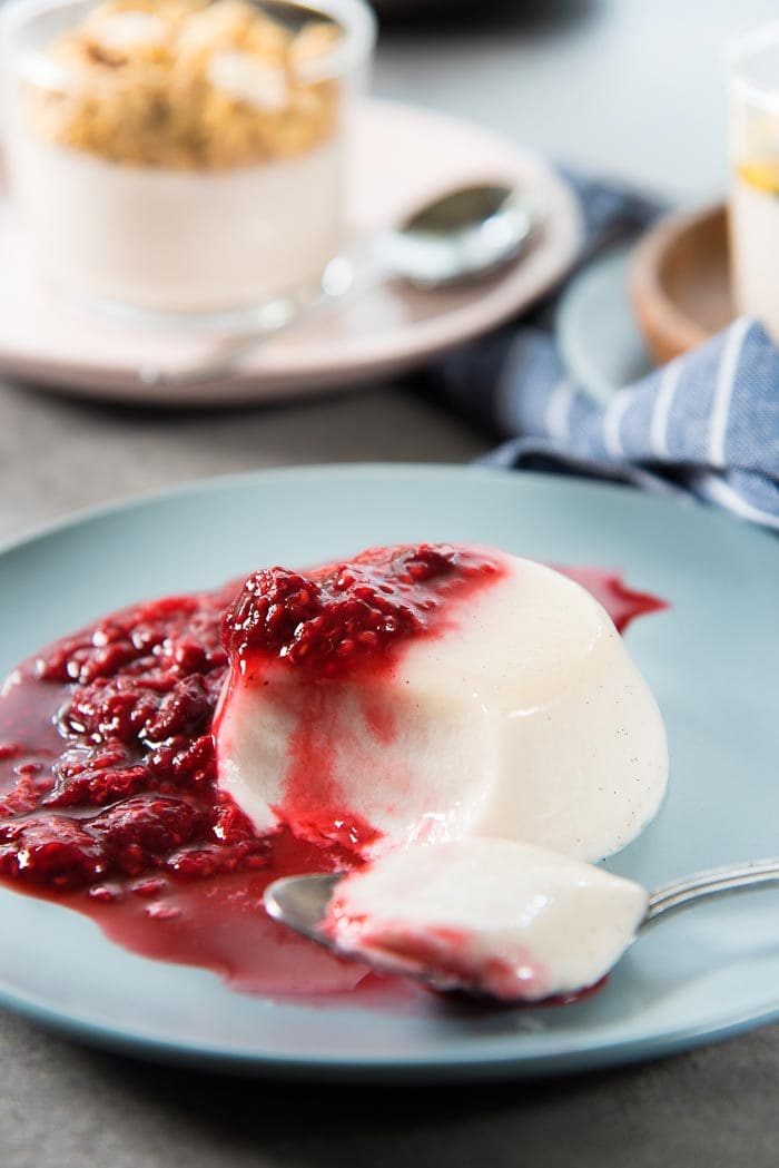 Paleo coconut panna cotta with raspberries on a blue plate, with a spoonful of the panna cotta placed in front. The texture of the panna cotta is smooth and silky and extremely soft too. 