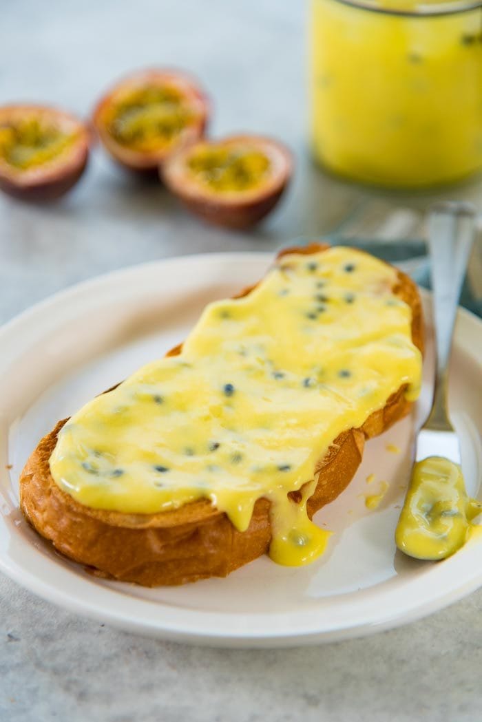 A thin layer of passion fruit curd spread on a piece of toast, and served on a white plate. This is one of the ways to use Passion fruit curd. 