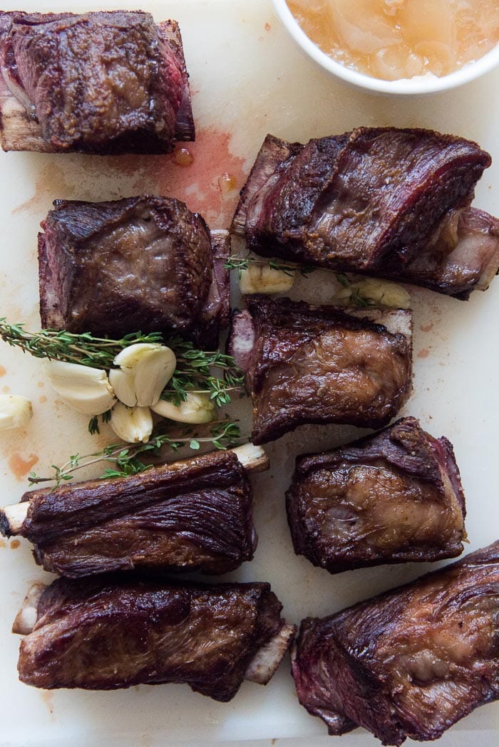 Short Ribs that have been seared and caramelized on a white board with whole garlic cloves and thyme. 