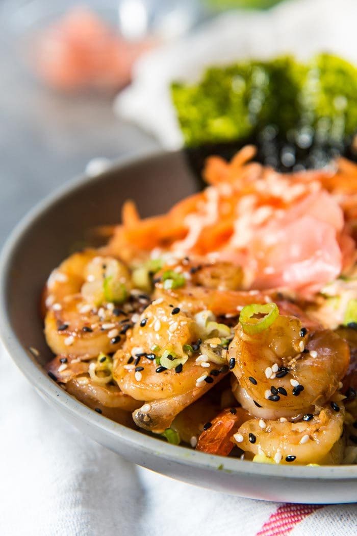 A close up of the teriyaki shrimp with sesame seeds, on top of Jasmine rice and other additions such as nori sheets. 