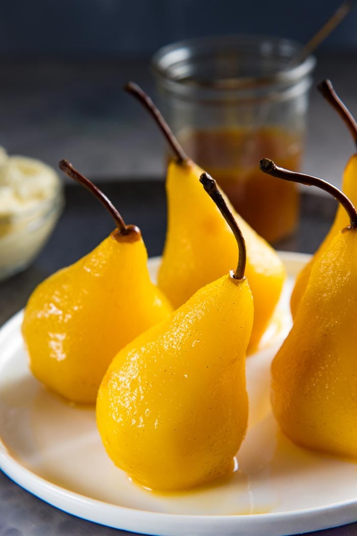 Five perfectly poached pears in saffron, cardamom and orange, on a white plate, on a dark tray with the saffron syrup and whipped mascarpone