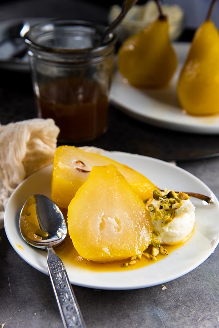 A saffron poached pear cut in half on a small white plate, placed on the saffron syrup. Whipped mascarpone cheese with chopped pistachios on the side. 