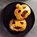 Two Halloween-themed, golden brown chorizo hand pies on a black plate, where the pastries feature smiling, spooky faces.