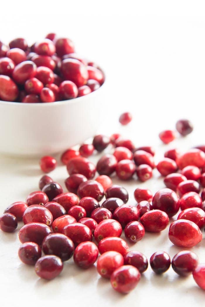 Fresh Cranberries in a bowl, with some on a white table top in the foreground. 