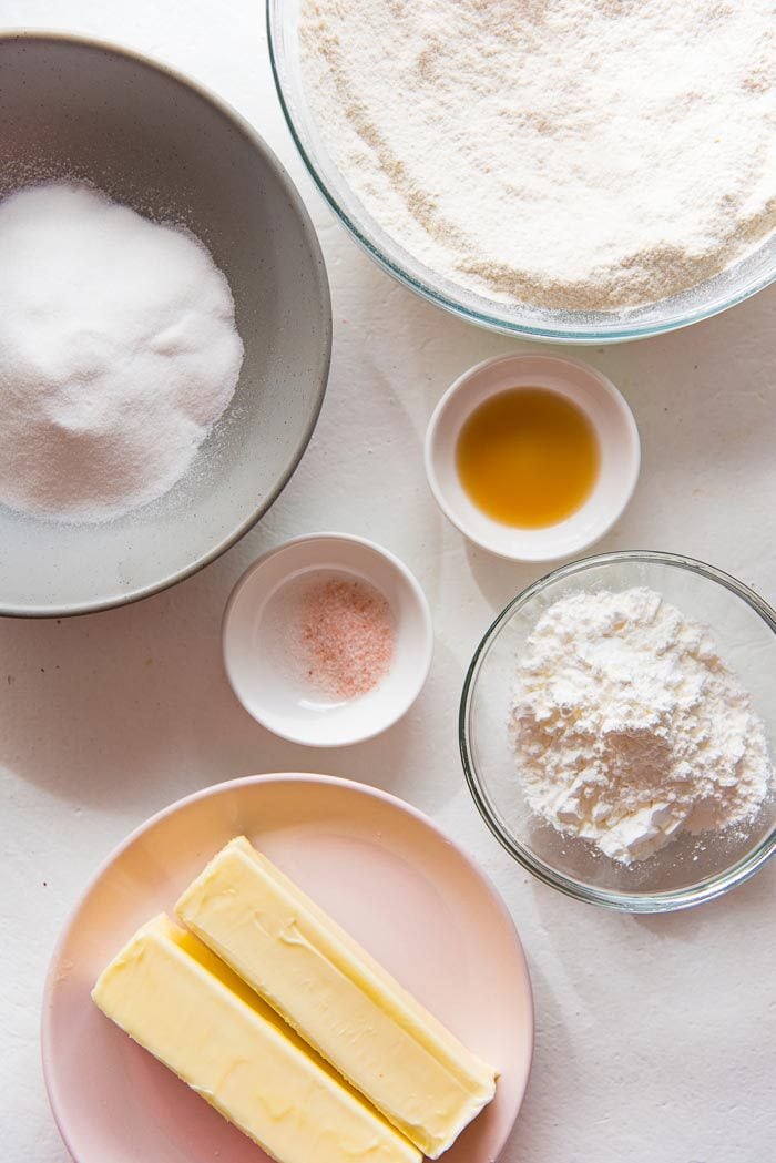 Ingredients for thumbprint cookies on a white table. Softened butter, sugar, white sugar, vanilla, salt, and flour in separate bowls and plates. 