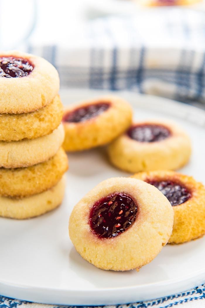 Classic thumbprint cookies placed on a white plate, with four cookies on a plate and a stack of cookies behind them.