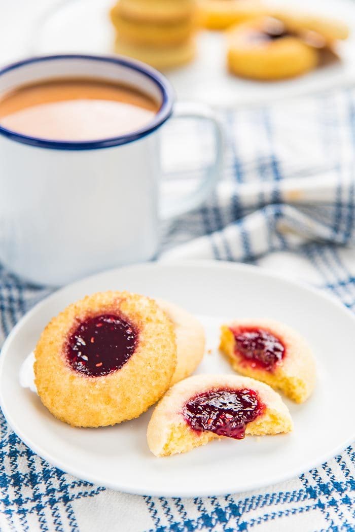 Three thumbprint cookies on a small white plate, with a mug of coffee in the background. The thumbprint cookie in the foreground broken in half. 