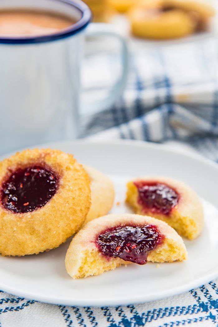 Thumbprint cookies with raspberry jam in the filling, on a white plate, with one broken in half, and the other behind it. A coffee mug and more thumbprint cookies int he background. 