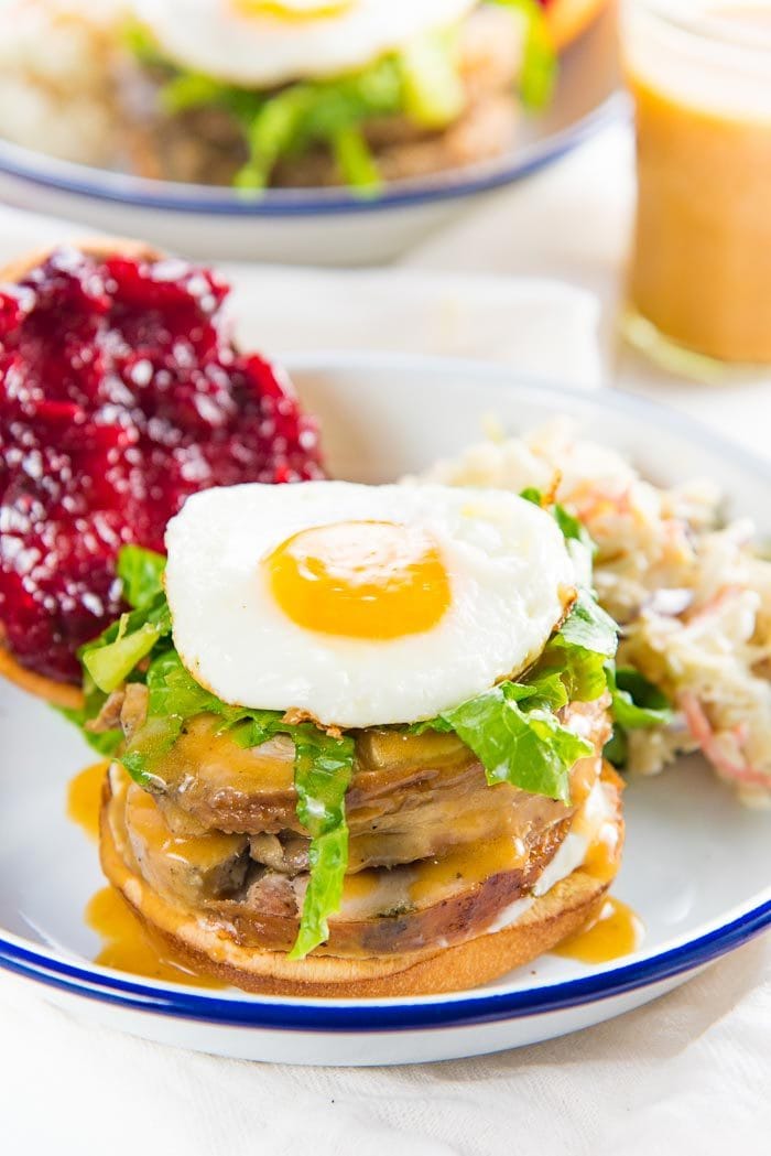 A leftover turkey sandwich on a white plate, topped with turkey, gravy, lettuce and an egg. The cranberry sauce spread bun and coleslaw in the background.