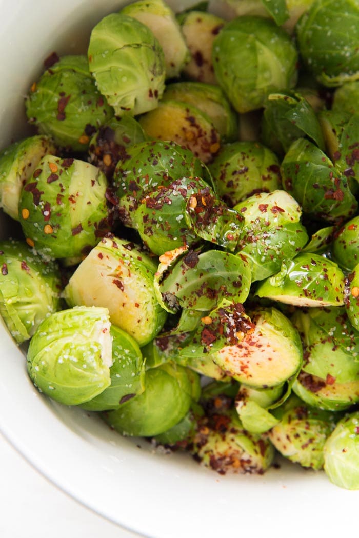 Halved Brussels sprouts in a white bowl, with chili flakes, salt and maple syrup added.