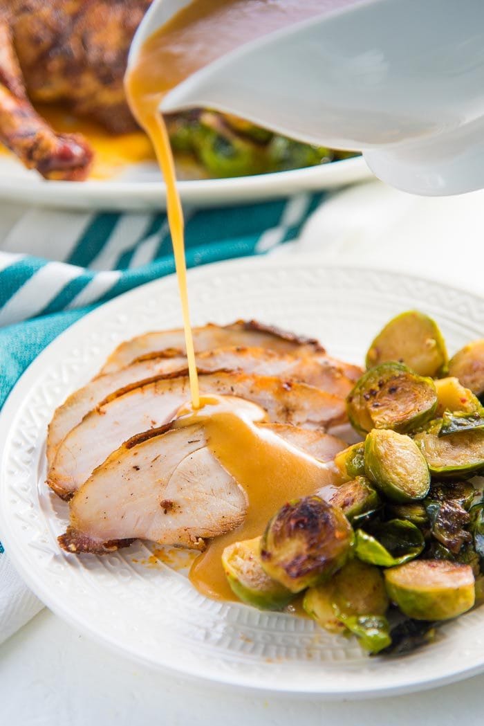 A plate of sliced oven roasted turkey breast slices on a plate, with gravy being poured over the turkey slices, on a white plate. Brussels sprouts as a side dish.