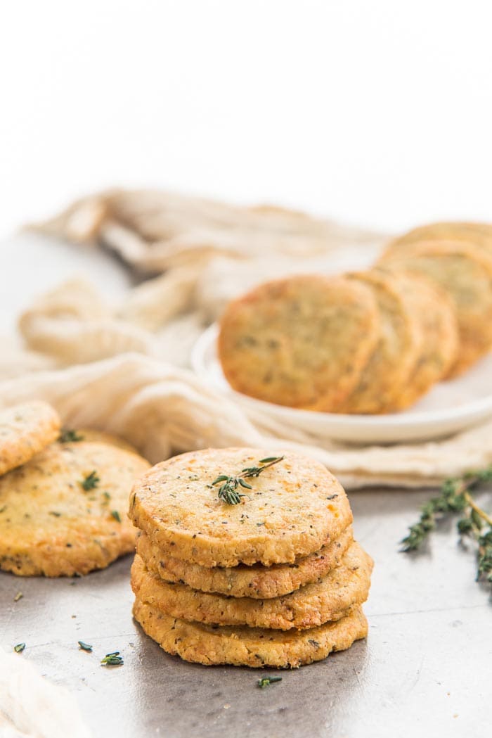 A stack of thyme and cheddar cheese cookies placed in the foreground with more cheddar cookies in the background on a white platter, with sprigs of thyme. 