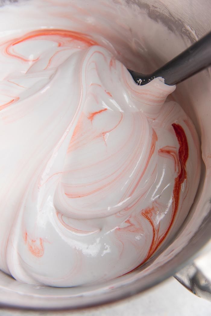 A metal mixing bowl with white marshmallow base with red swirls, and a silicone spatula