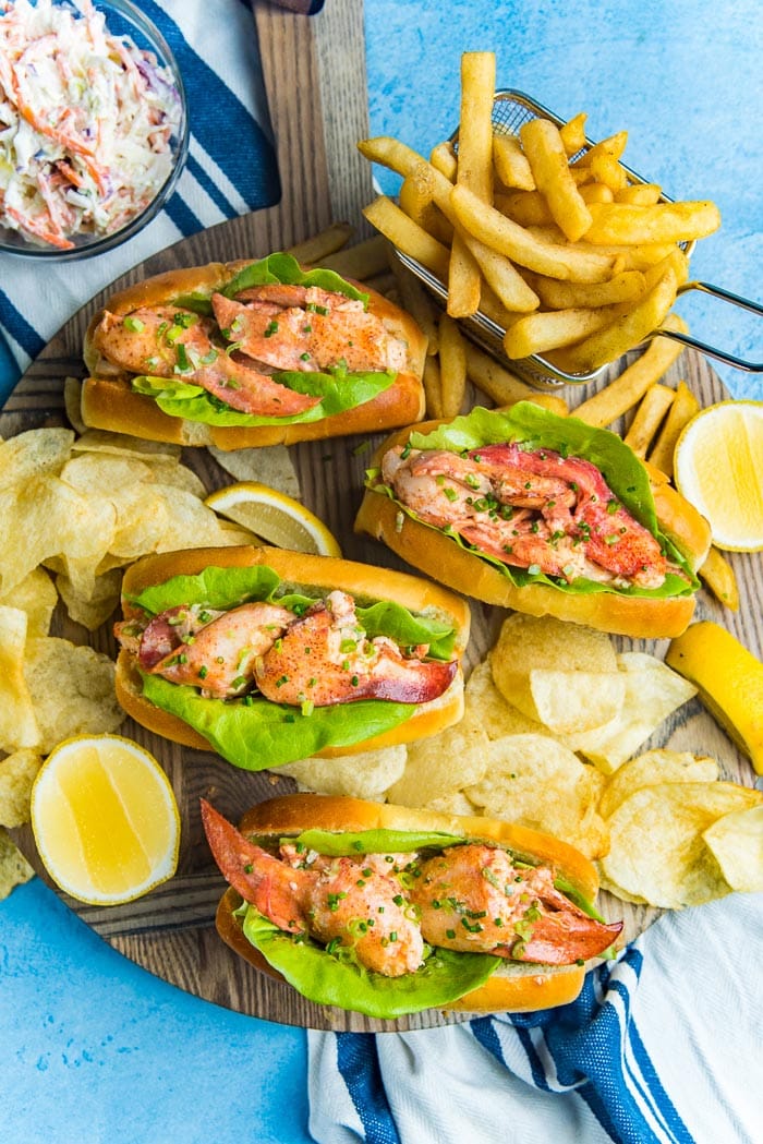 An overhead view of Maine lobster rolls on a wooden serving board with potato chips and french fries