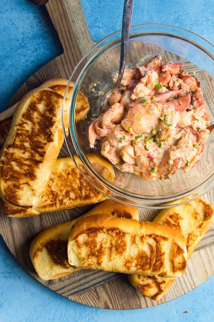 Toasted top split buns (or lobster roll buns) on a wooden board with a bowl of lobster for lobster rolls.