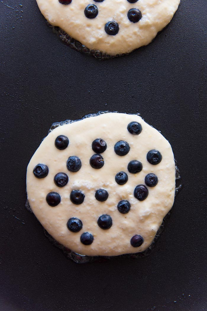 Blueberry pancakes being cooked on a griddle with blueberries placed on top. 